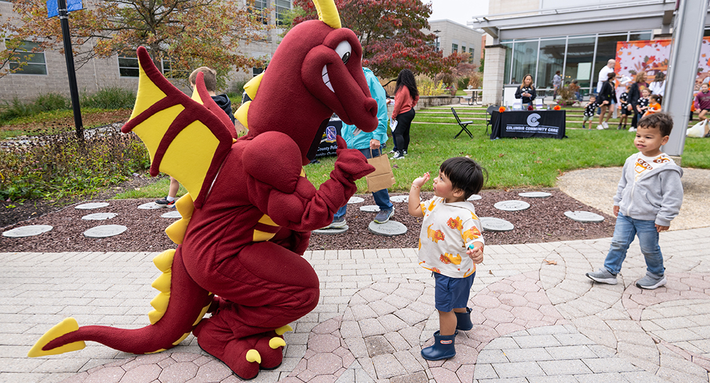 HCC’s red dragon mascot Duncan crouches to greet a young child on a paved campus walkway during an outdoor fall family event, while another child watches nearby.