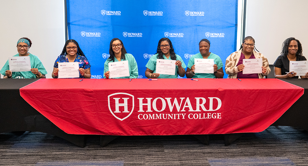 Seven women sit behind a table draped with a red Howard Community College banner, each holding a certificate and smiling in front of a blue HCC branded backdrop during an Apprenticeship Week signing event.