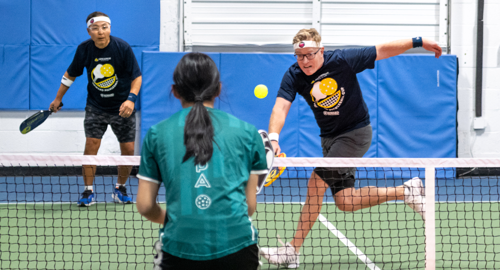 Two pickleball players on an indoor court compete at the net, with one player lunging forward to hit the ball while their partner stands ready behind them. An opposing player in a green shirt faces them from across the net.