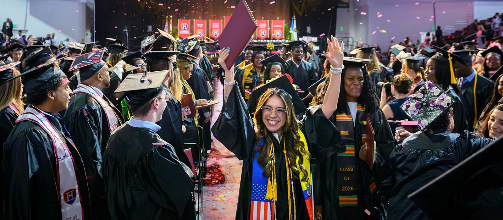 HCC graduates celebrate during commencement ceremony, raising diplomas and cheering amid confetti as students in caps and gowns fill the arena.