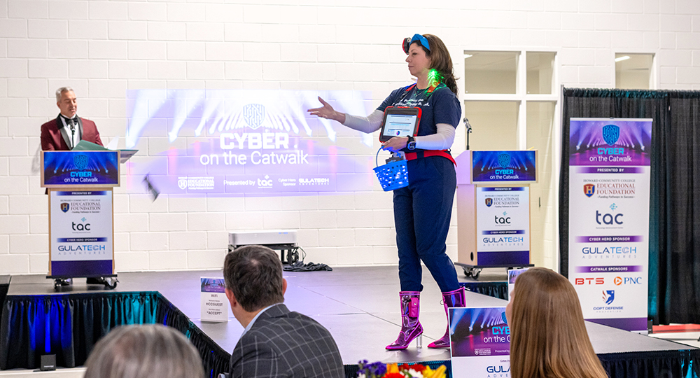 Presenter wearing a glowing, cyber-themed outfit walks down a runway during the “Cyber on the Catwalk” event while a host speaks at a podium and audience members watch.