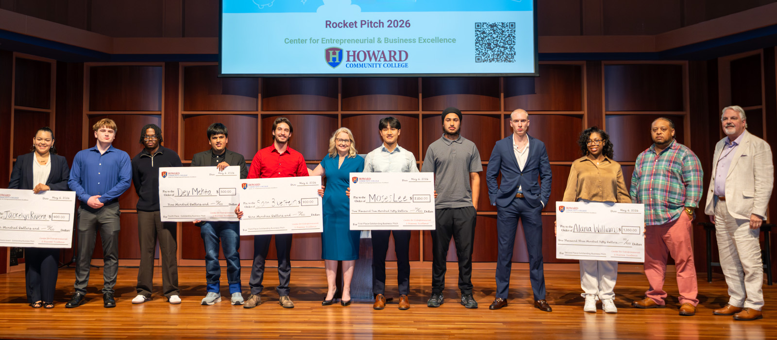 Group photo of Howard Community College Rocket Pitch 2026 participants and organizers standing on stage with oversized award checks beneath a presentation screen displaying the event title and college logo.