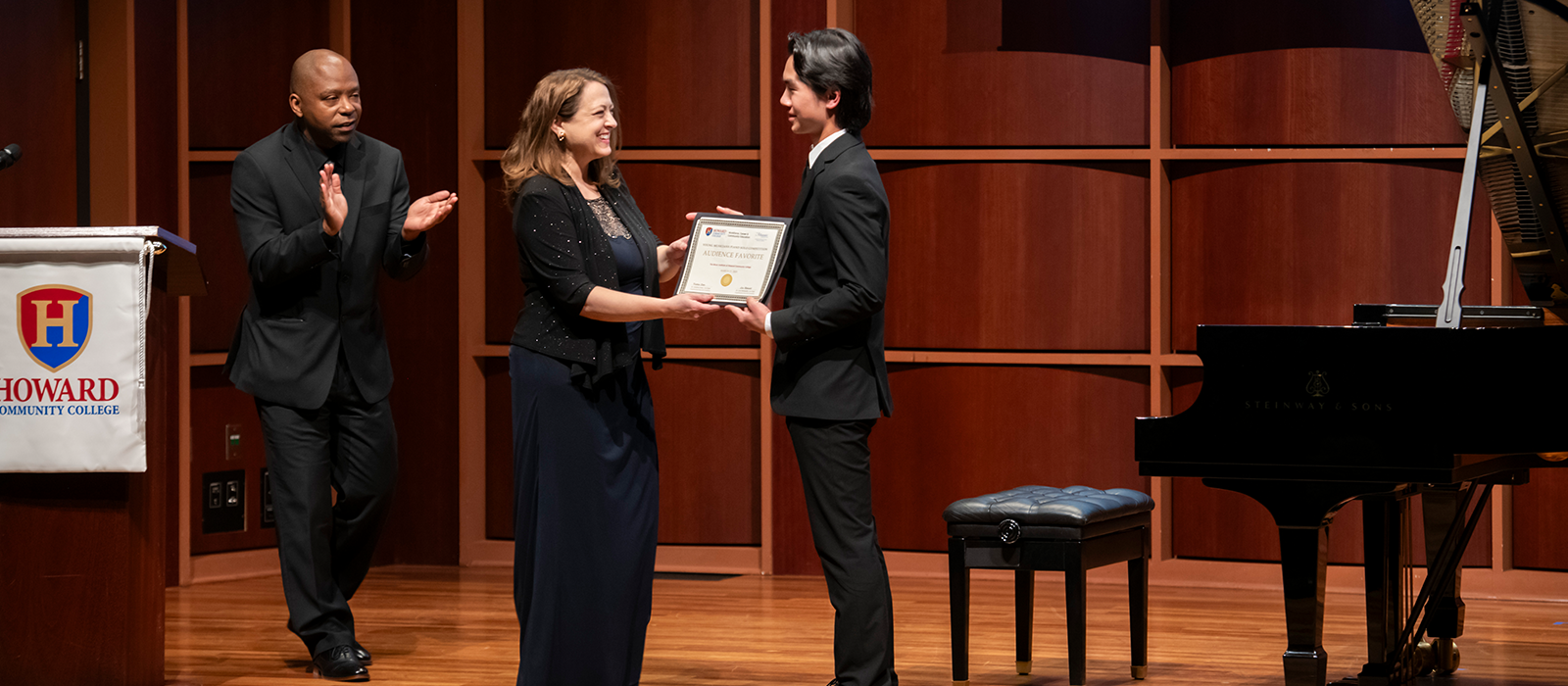 Student musician receives an audience award on stage at a concert hall, with presenters applauding beside a Steinway grand piano.
