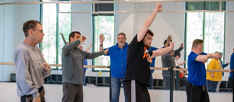 Participants raise their hands during a group fitness class in a bright studio space, with other participants exercising in the background.