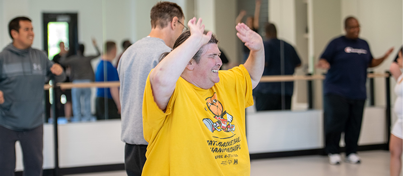 Student in a yellow T-shirt raises their hands and smiles during a group fitness class in a bright studio space, with other students exercising in the background.