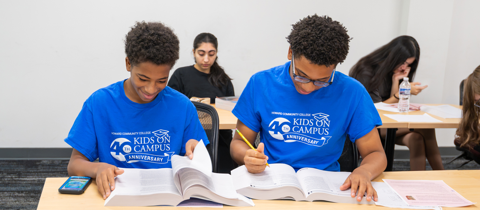 Two students in Kids on Campus T-shirts work on assignments at a classroom table while other students study in the background.