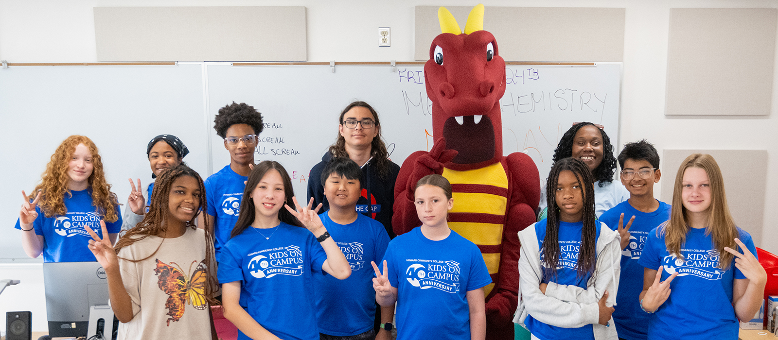 A group of young students in Kids on Campus T-shirts pose with the HCC dragon mascot in a classroom.