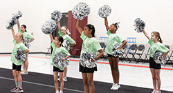Group of young cheerleaders performing indoors, standing in formation with silver pom-poms raised, wearing matching light green Kids on Campus shirts and black shorts.