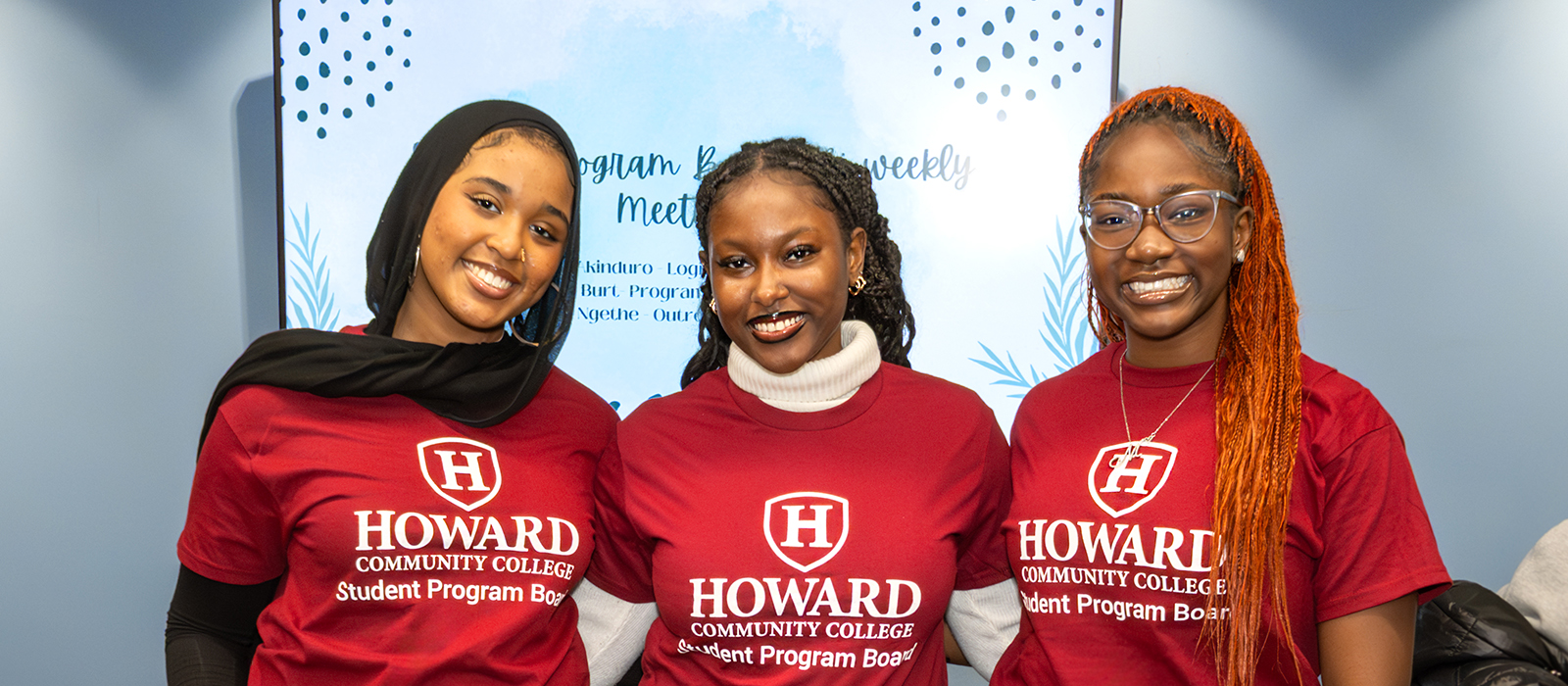 Three smiling Howard Community College students stand together in front of a blue patterned backdrop. They are wearing matching red “HCC Student Program Board” T-shirts. The student on the left wears a black hijab, the student in the middle has braided hair and a white turtleneck under her shirt, and the student on the right has long orange braids and glasses.